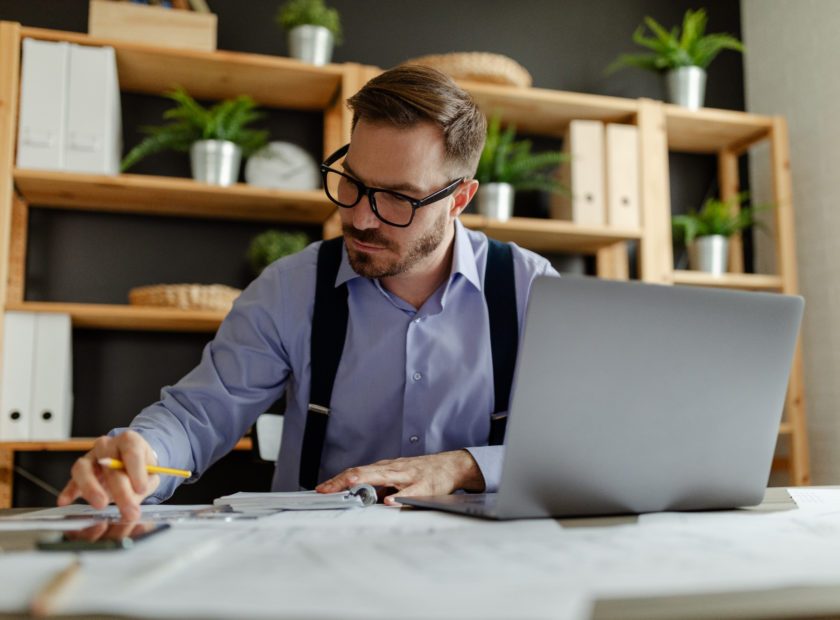 Man with eyeglasses reading information from paper documents and checking in database on laptop.