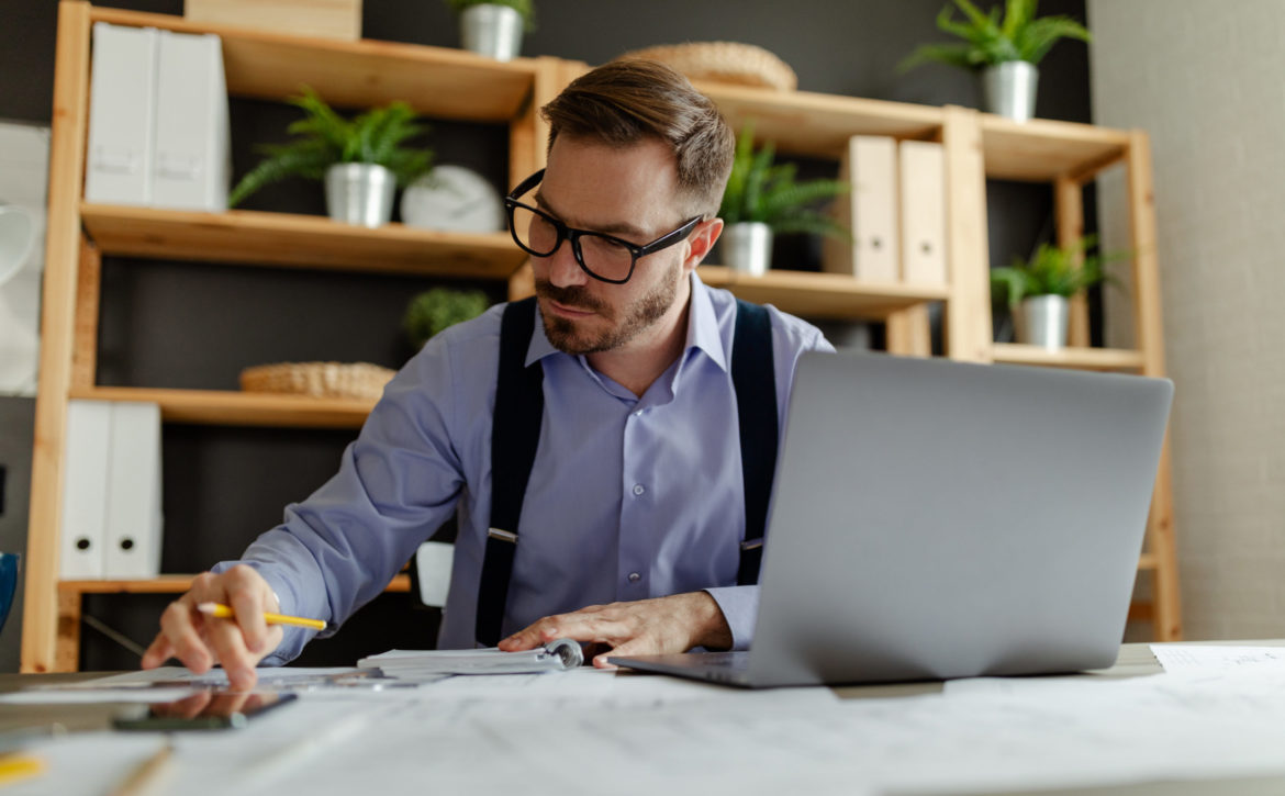 Man with eyeglasses reading information from paper documents and checking in database on laptop.