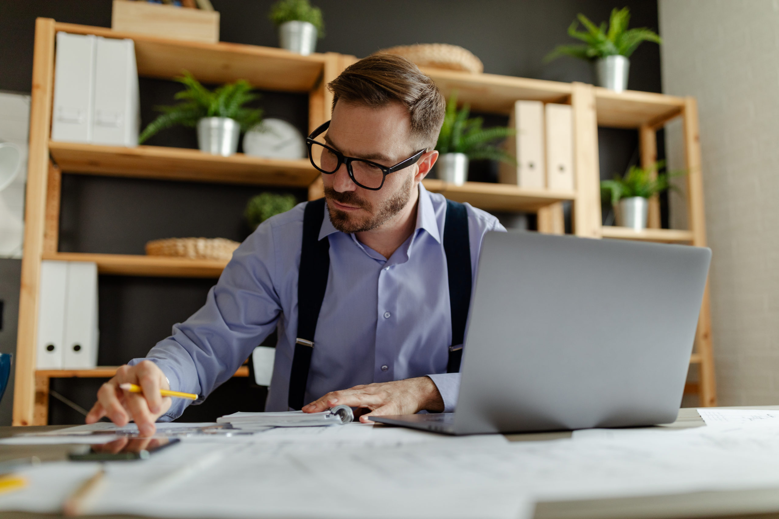 Man with eyeglasses reading information from paper documents and checking in database on laptop.